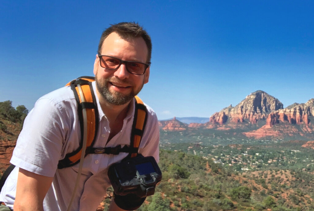 An image of Michael from atop a mountain. He wears a hiker's backpack with a camera fastened to his front. In the background a green valley stretches to the distance, punctuated by red mountains.